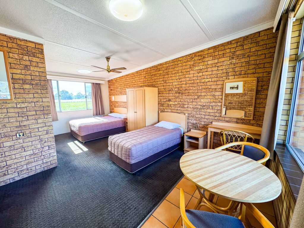 A wide-angle view of Nandewar Motor Inn's room with two single or double beds, both covered with dusty-rose or mauve quilted comforters and white pillows.