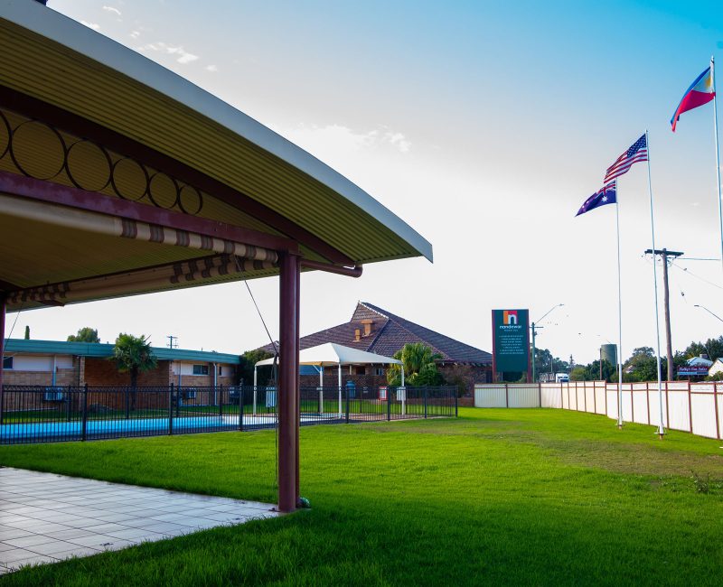 A wide view of a motel property with a foreground dominated by a bright green lawn and a partially visible covered patio with a light-colored roof supported by dark red-brown posts.