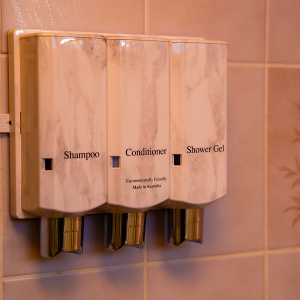 A close-up of a three-chamber, wall-mounted dispenser for bath products labeled "Shampoo," "Conditioner," and "Shower Gel."