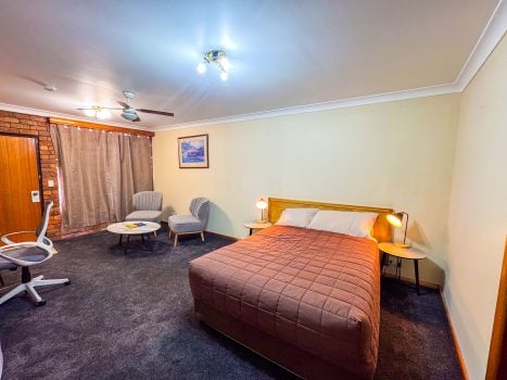 A wide-angle view of Nandewar Motor Inn room with a bed in the foreground covered by a dusty-rose or mauve quilted bedspread.