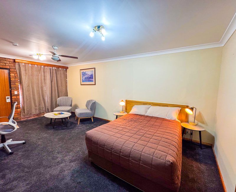 A wide-angle view of Nandewar Motor Inn room with a bed in the foreground covered by a dusty-rose or mauve quilted bedspread.
