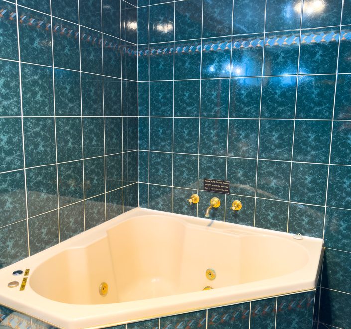 Nandewar Motor Inn's triangular corner spa tub with an off-white or cream-colored basin is set against walls covered in glossy, dark blue-green marbled square tiles with white grout.