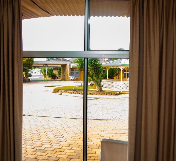 A view of Nandewar Motor Inn exterior courtyard seen through a large window with a vertical divider, framed on both sides by open, heavy beige curtains.