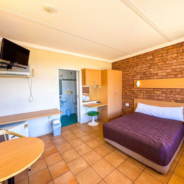 A large bed with a dark purple quilted comforter and white pillow is situated on the right against a wall covered in red-brown brick veneer.