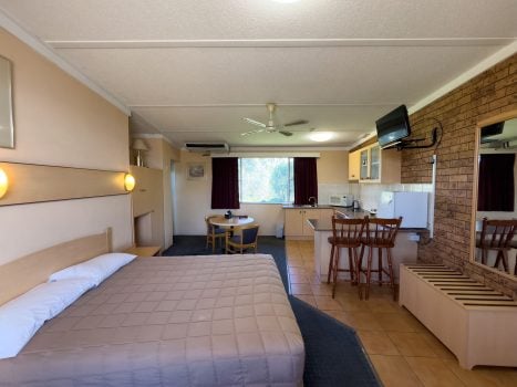 A spacious motel room interior with a double or queen-sized bed in the foreground, covered with a light brown/tan quilted comforter.