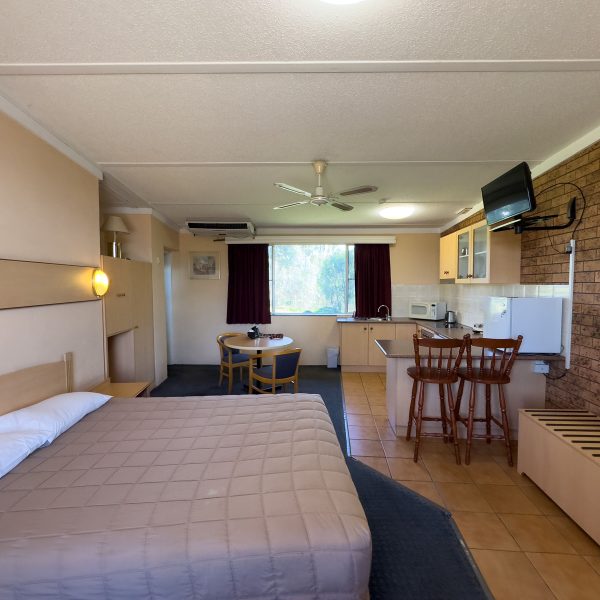 A spacious motel room interior with a double or queen-sized bed in the foreground, covered with a light brown/tan quilted comforter.