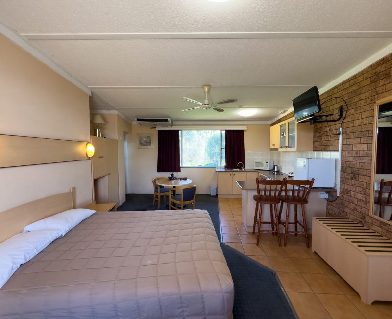 A spacious motel room interior with a double or queen-sized bed in the foreground, covered with a light brown/tan quilted comforter.