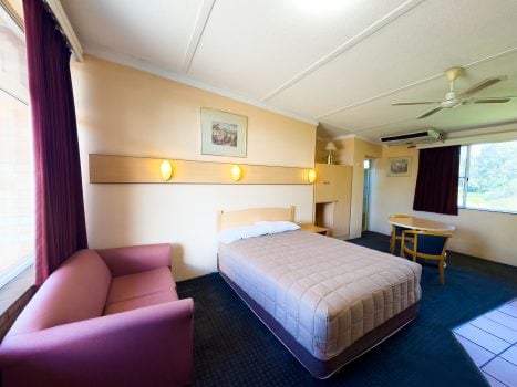 A Nandewar Motor Inn room featuring a double or queen-sized bed in the center, covered with a light brown or tan quilted comforter and white pillows, set against a light wood headboard.