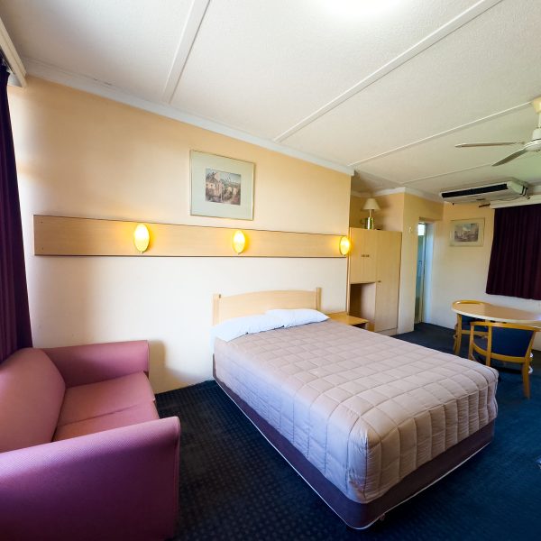 A Nandewar Motor Inn room featuring a double or queen-sized bed in the center, covered with a light brown or tan quilted comforter and white pillows, set against a light wood headboard.