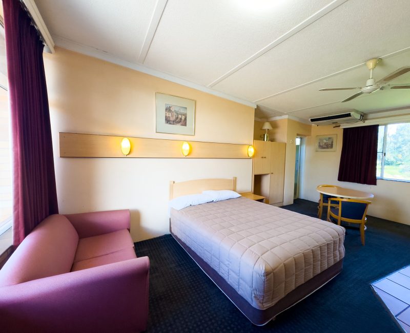 A Nandewar Motor Inn room featuring a double or queen-sized bed in the center, covered with a light brown or tan quilted comforter and white pillows, set against a light wood headboard.