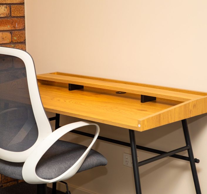 A light wood desk with a raised monitor shelf and black metal A-frame legs is positioned against a plain light beige wall.