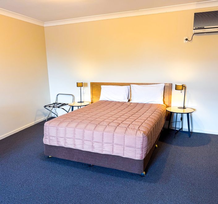 A double or queen-sized bed dominates the center of the frame, covered with a dusty-rose or mauve quilted comforter and white pillows, and set against a light-toned wooden headboard.